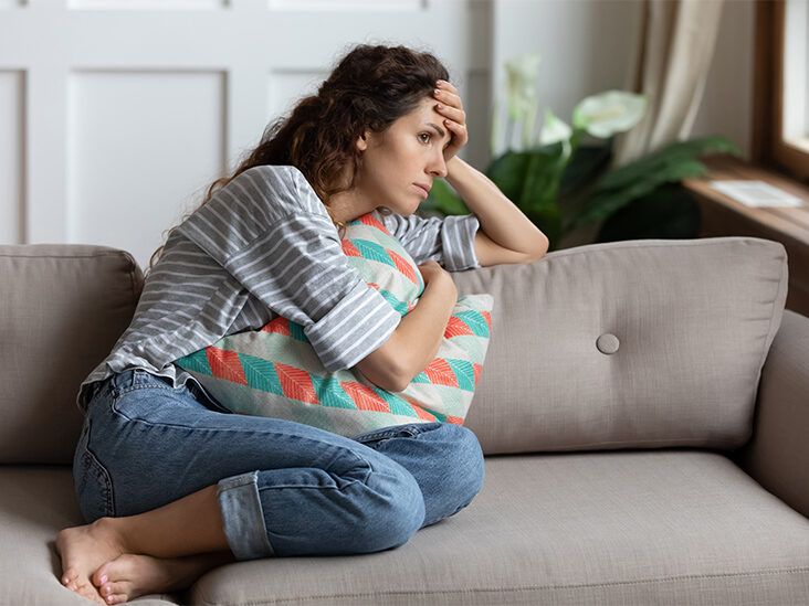 Woman sitting on a couch, holding a colorful pillow, displaying a contemplative and distressed expression, reflecting emotional burden related to past mistakes and the impact of a criminal record.