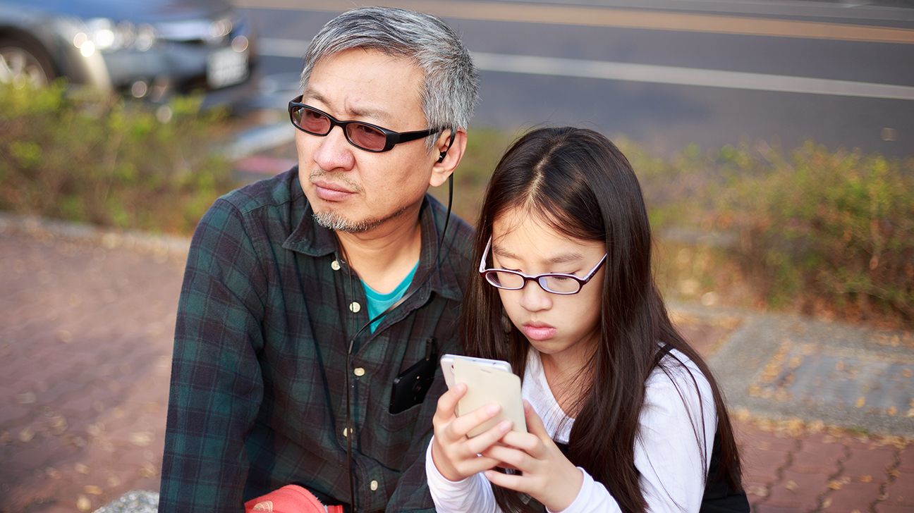a parent wearing glasses sits outside with their child, also in glasses, who is looking at a phone