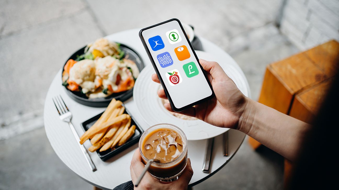 Person eating a meal while holding their phone. The phone screen shows logos for calorie counter apps. 