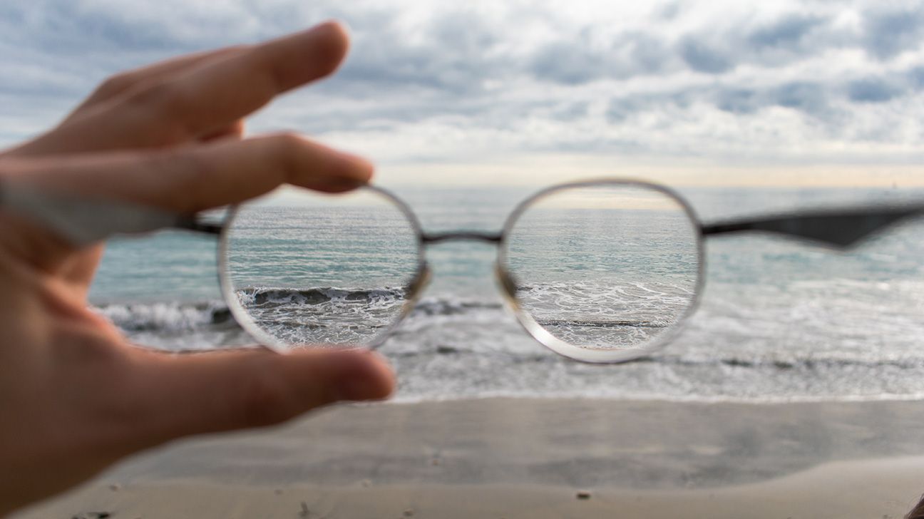 Someone holding their glasses out, showing us a small bit of the ocean in focus.
