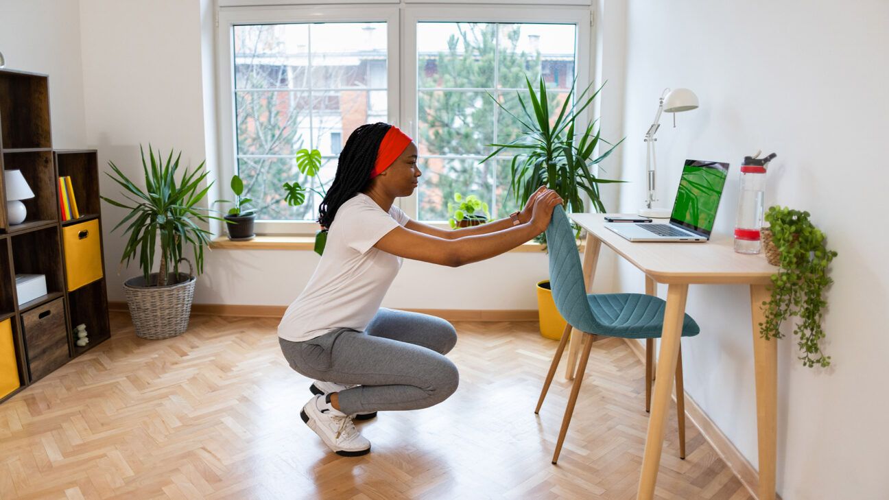 A woman does squats next to a desk.
