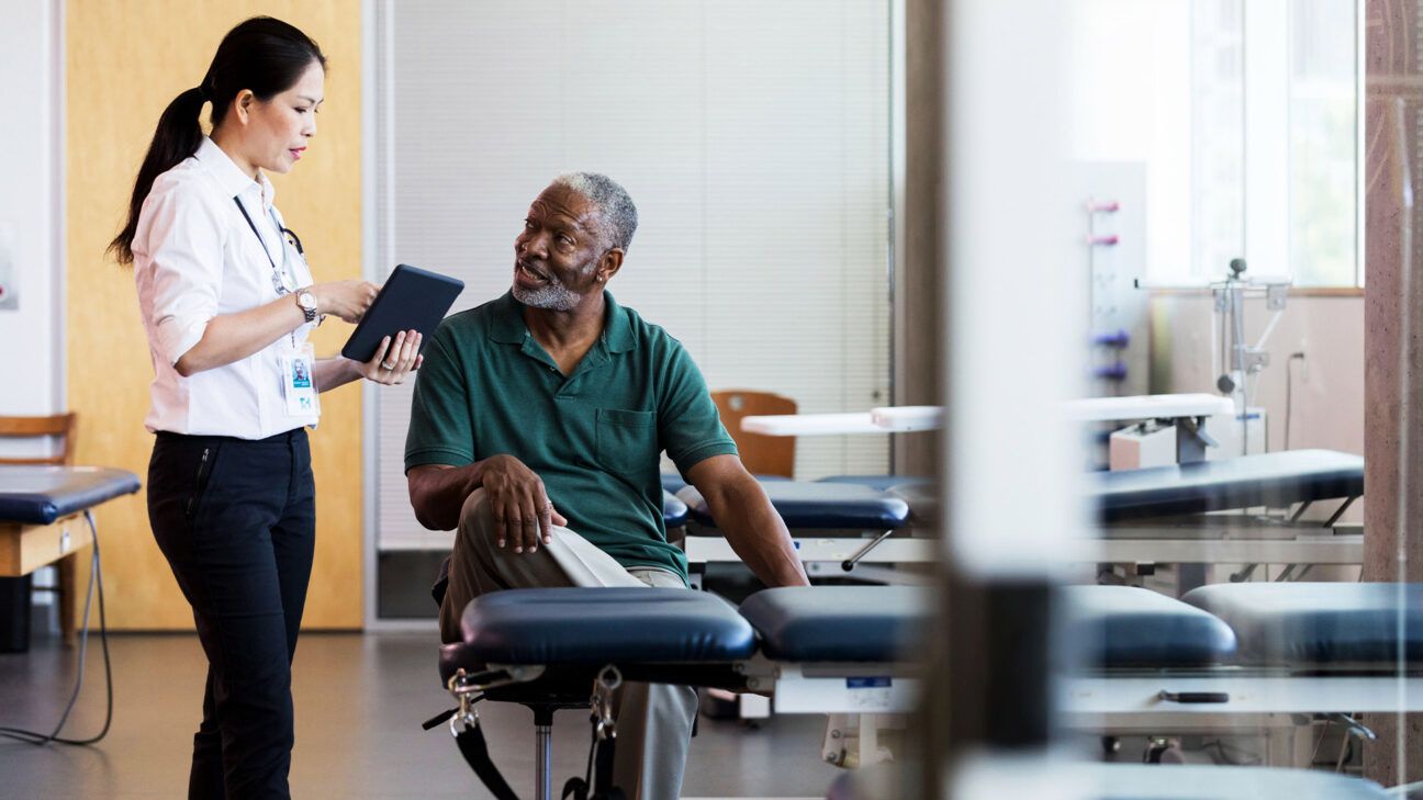 A man in green shirt talks to physician in white shirt and black pants.