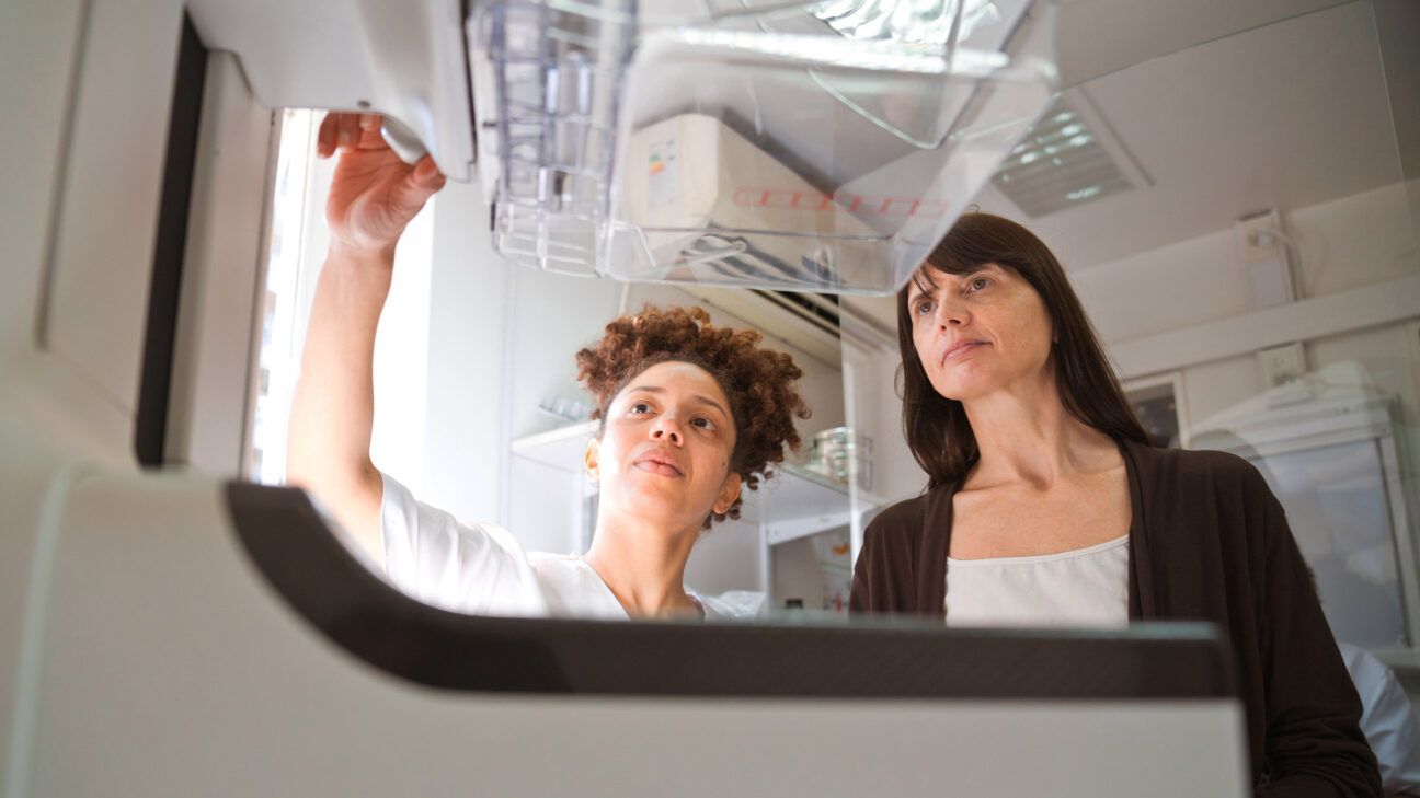 A healthcare provider in scrubs explains a mammogram machine to a woman in a dark sweater.