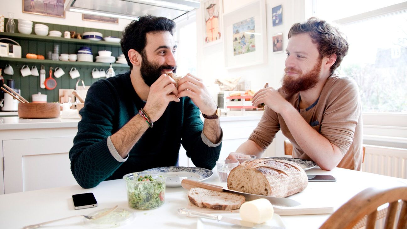 Two men with beards sit at a table eating sandwiches.