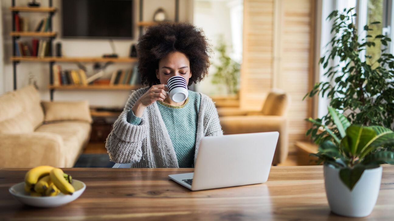 Woman in grey robe drinks from a mug in front of a laptop.