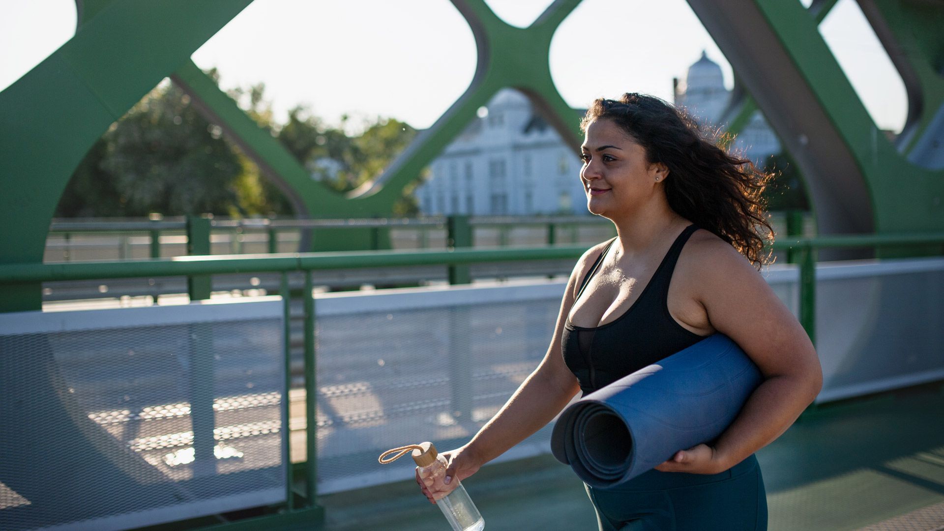 Woman walking over city bridge with yoga mat and water bottle, going to workout. 