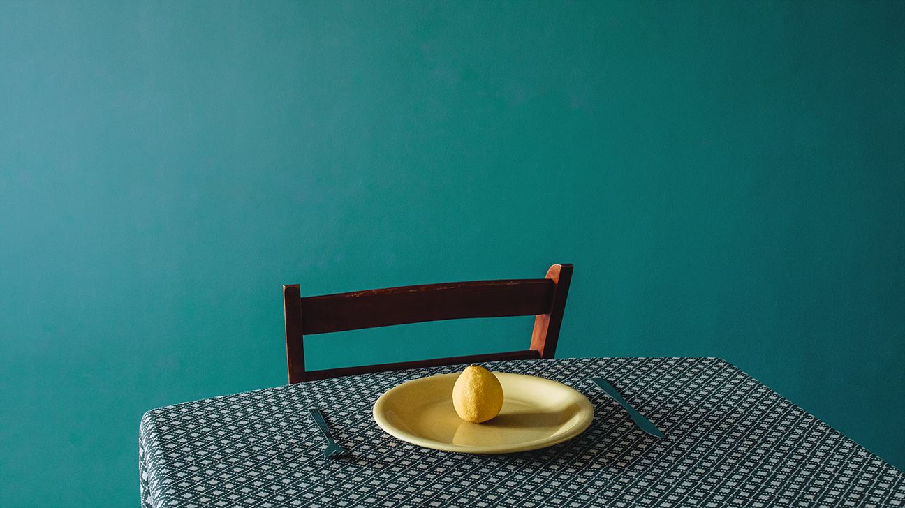 A sour lemon sitting alone on a yellow plate, offset by a bright blue wall. 