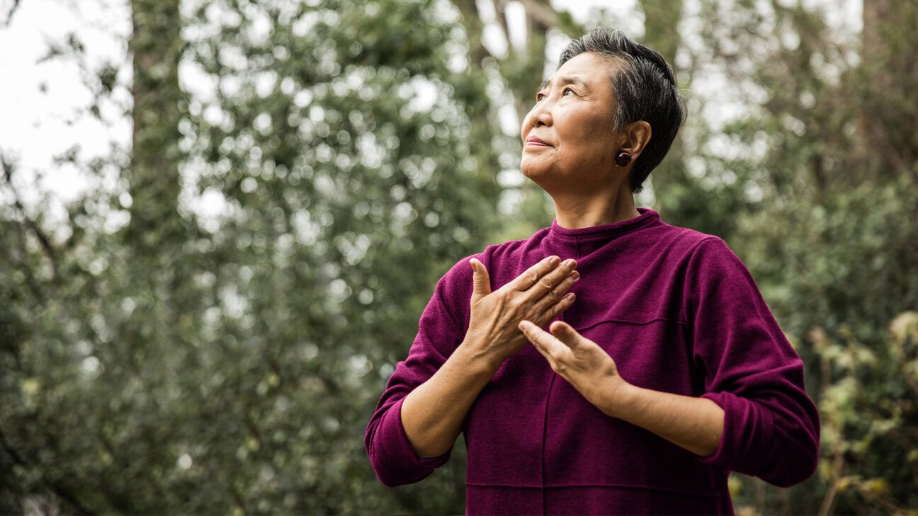 Woman in maroon top practices tai chi while outside.