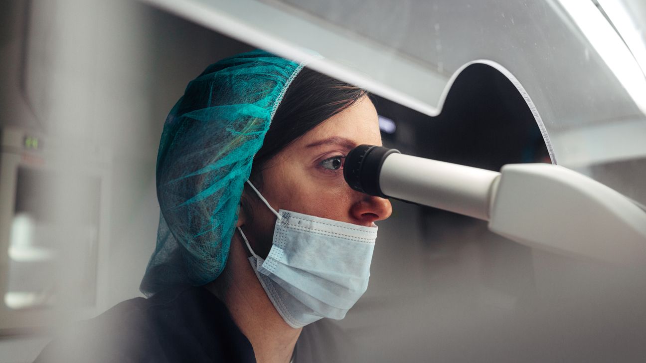 Woman in lab gear looks into microscope.