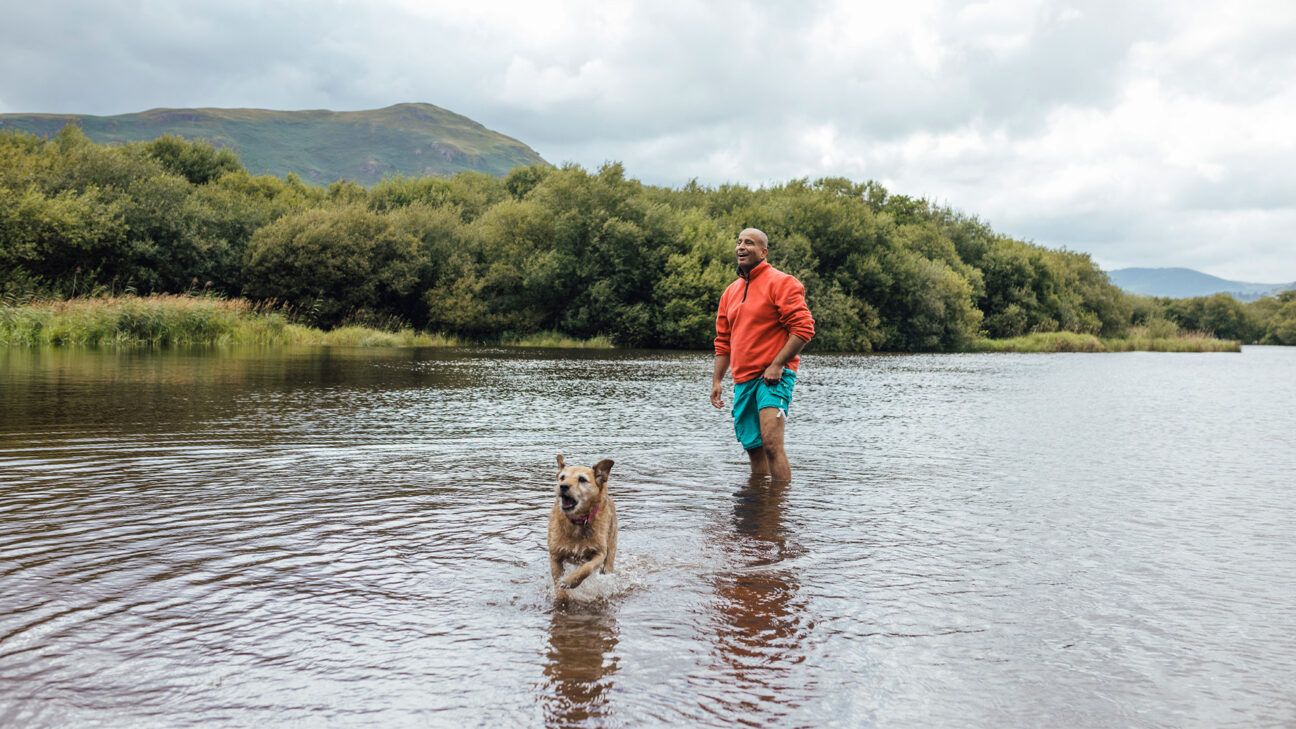 Man in orange shirt and blue shorts walks in a river with his dog.