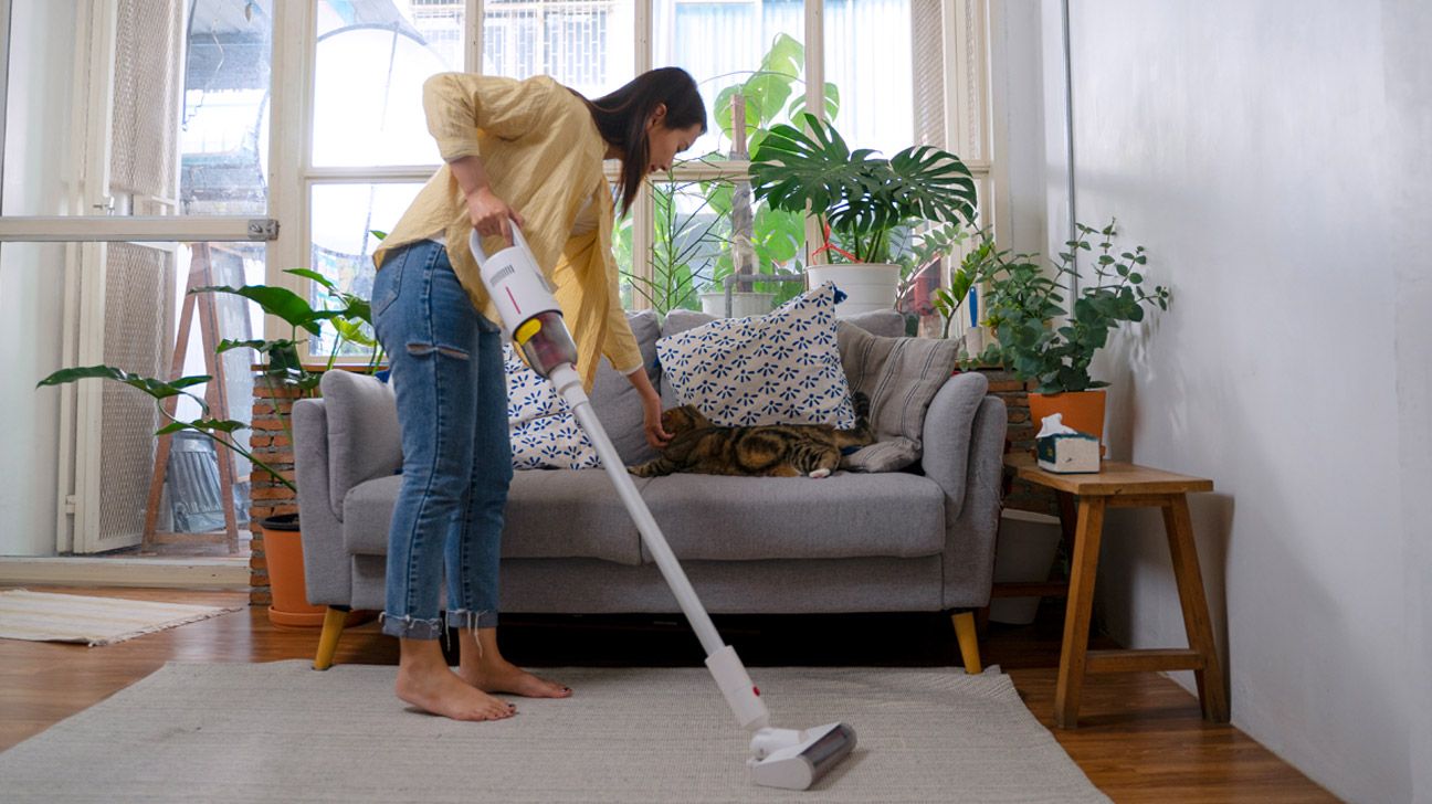 A white woman vacuuming her living room while she pets her cat.