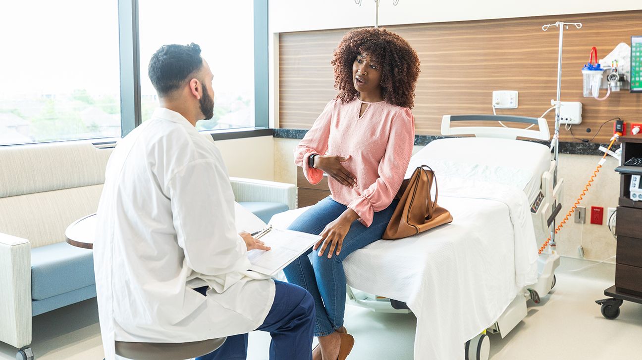 A black female wearing a pink top and jeans sat on the end of a hospital bed, speaking with a healthcare professional.