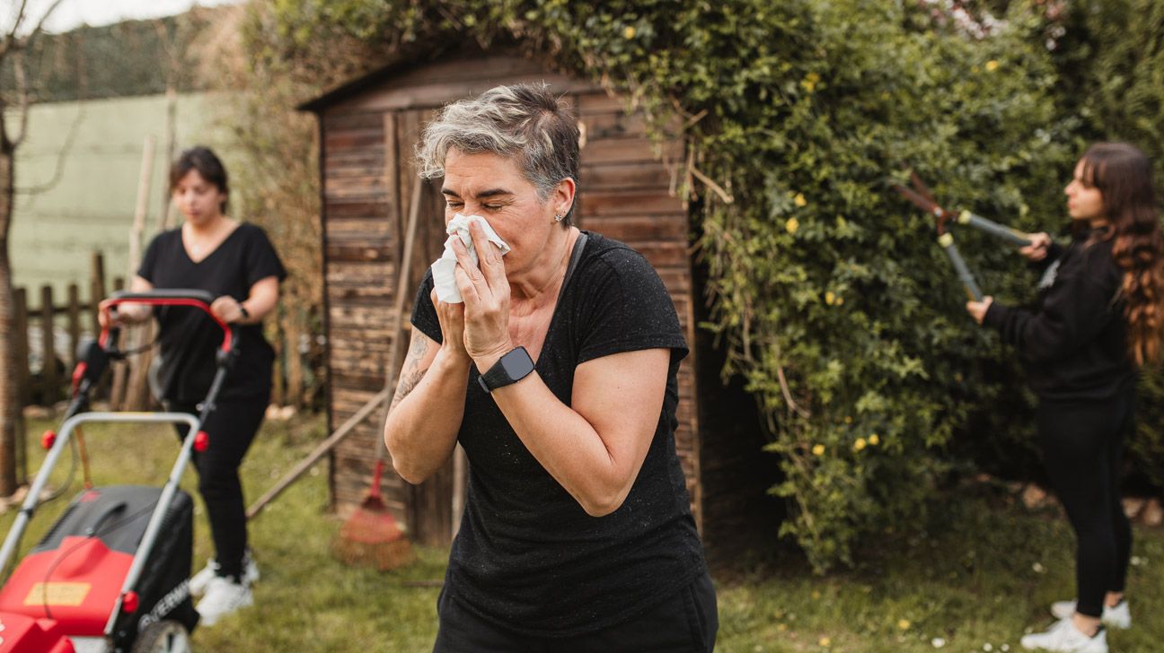 Woman with short, gray hair sneezing into a tissue outside while others do yard work