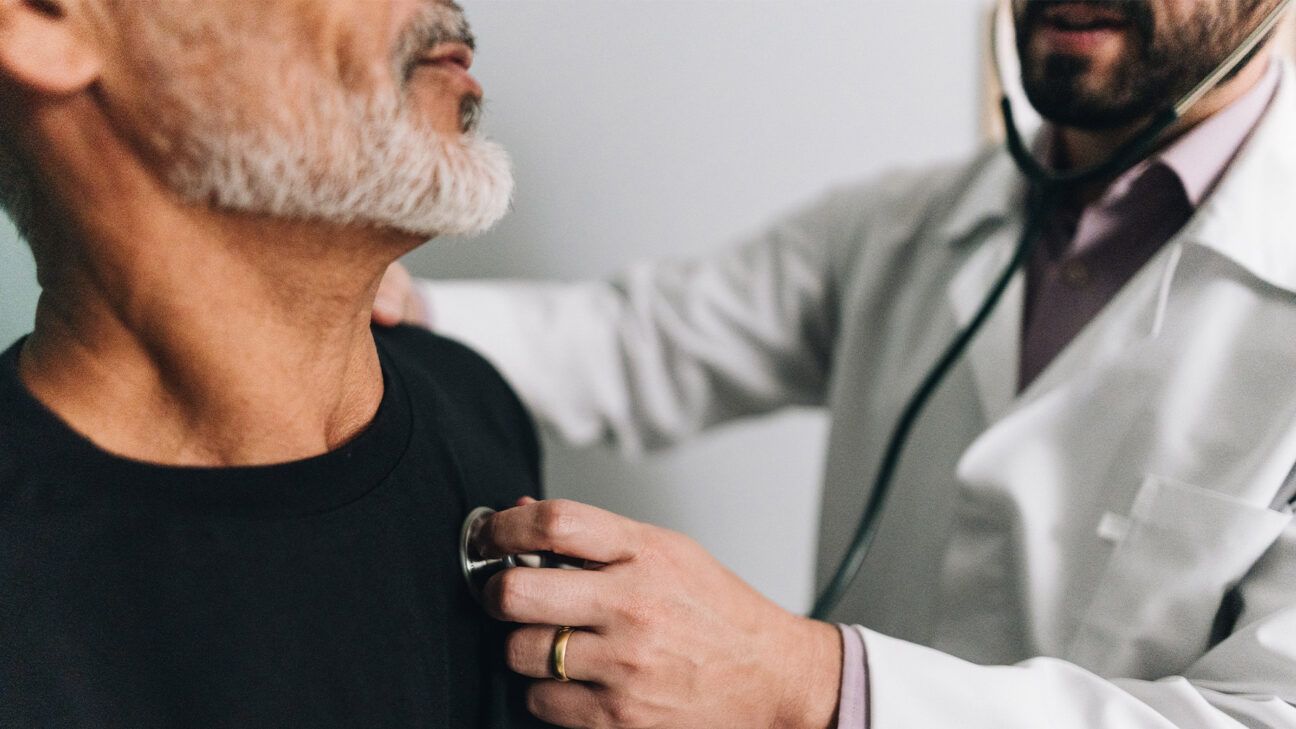 Physician uses stethoscope to listen to heart of man in black shirt.