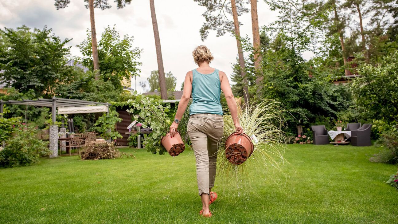 Woman seen from behind carries two potted plants in a yard.