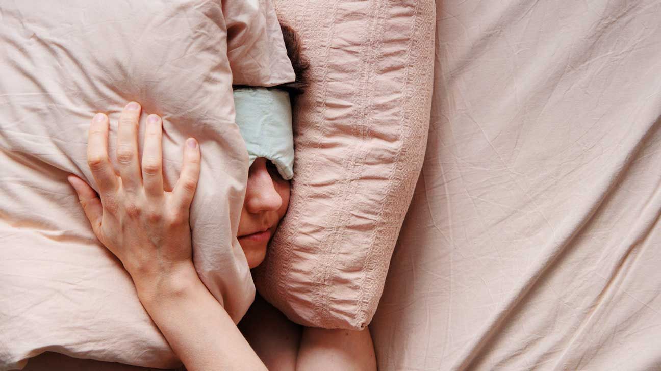 Person wearing an eye sleep mask while their head is placed between two pink pillows.