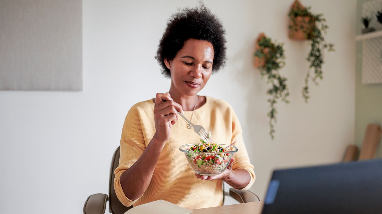 Adult Black woman eating quinoa salad while using laptop at home office