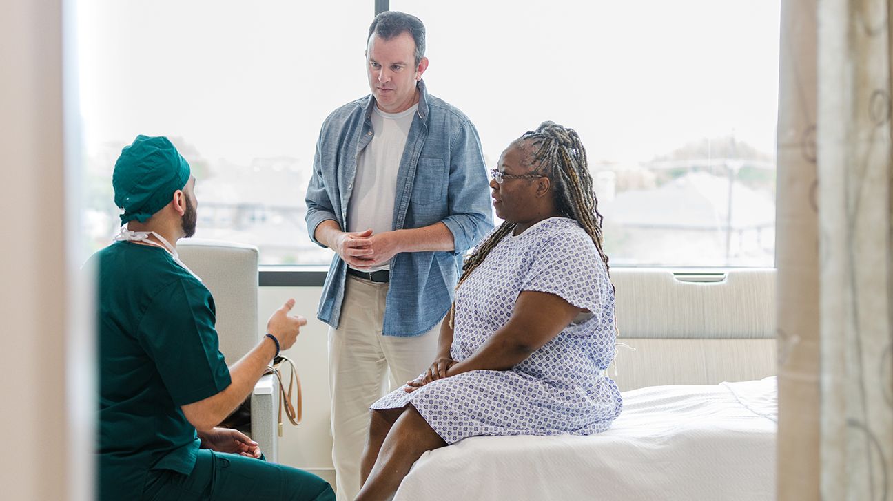 A patient wearing a hospital gown, sitting on a hospital bed, and talking with a surgeon.