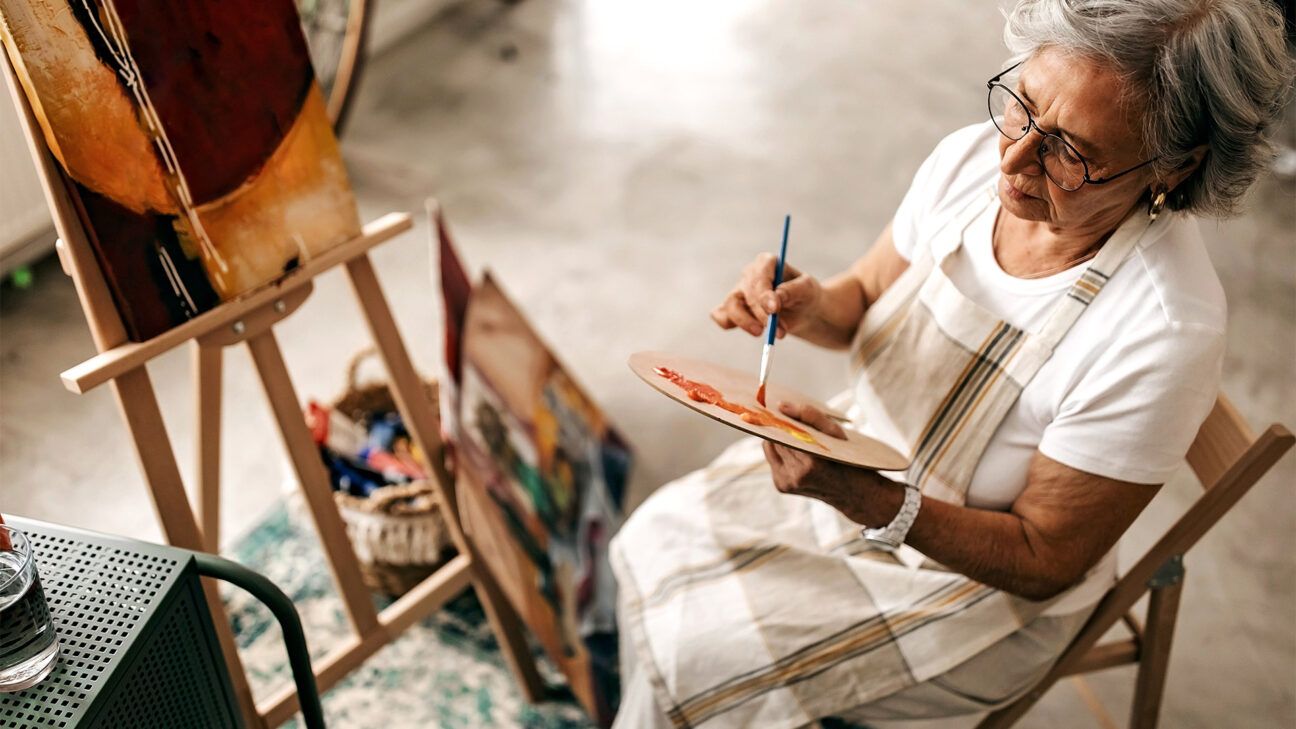 Woman in checkered dress with grey hair paints on an easel.