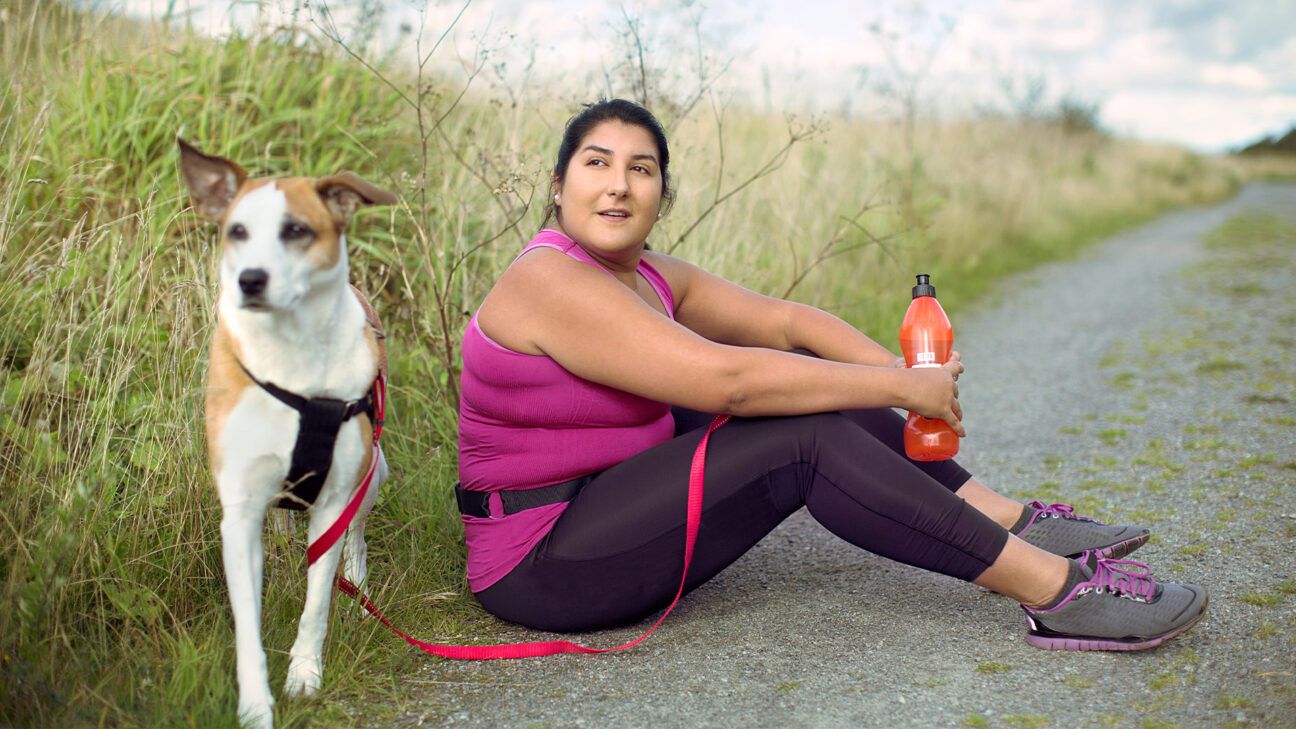 Woman in athletic wear takes a break on a road next to a white and brown dog.