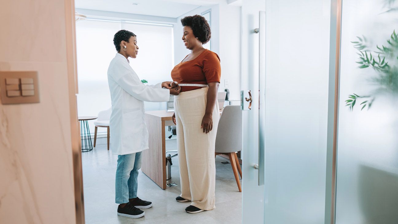 Scene viewed through an open door of two dark-skinned women with short, dark hair standing face-to-face in an office that has a small desk, some chairs, and a side table: the woman on the left in a lab coat is measuring the waist of the woman on the right (wearing street clothes) with a tape measure