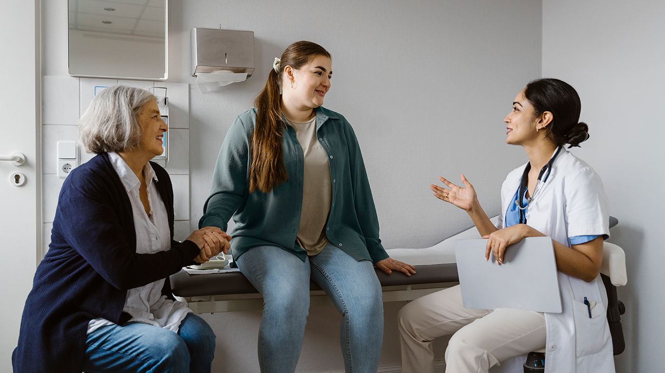 exam room setting with light-colored walls, towel holder and mirror: light-skinned female patient with long dark ponytail dressed in street clothes sitting on exam table looking to her left at doctor and smiling slightly, middle-aged light-skinned woman with short gray hair seated in a chair beside the patient wearing jeans and a light shirt and a dark cardigan holding the patient’s right hand in both of hers and smiling, light-skinned female with dark hair pulled back in a bun wearing a white lab coat and light-colored slacks and a stethoscope seated on other side of patient and holding a clipboard in left hand with right arm bent at elbow and right hand up and open and looking up at patient as if speaking