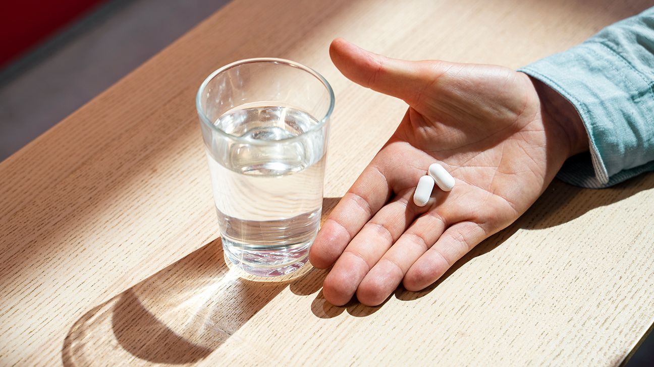 Two white pills in someone's hand next to water glass