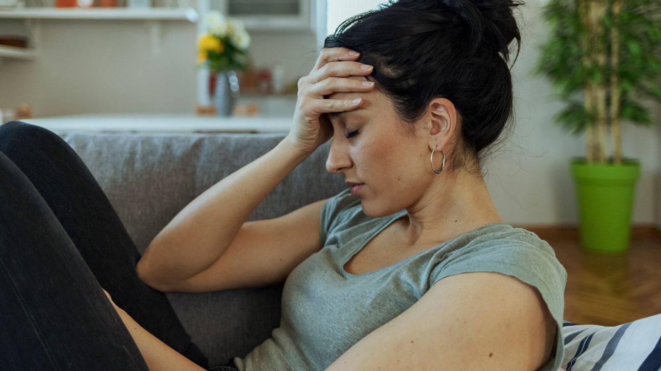 light-skinned woman with dark hair pulled back into a bun, wearing a light-colored T shirt and dark pants, on couch with knees bent and pulled toward her, eyes closed, left arm down with left hand at her lap, right elbow on right calf, right hand on forehead