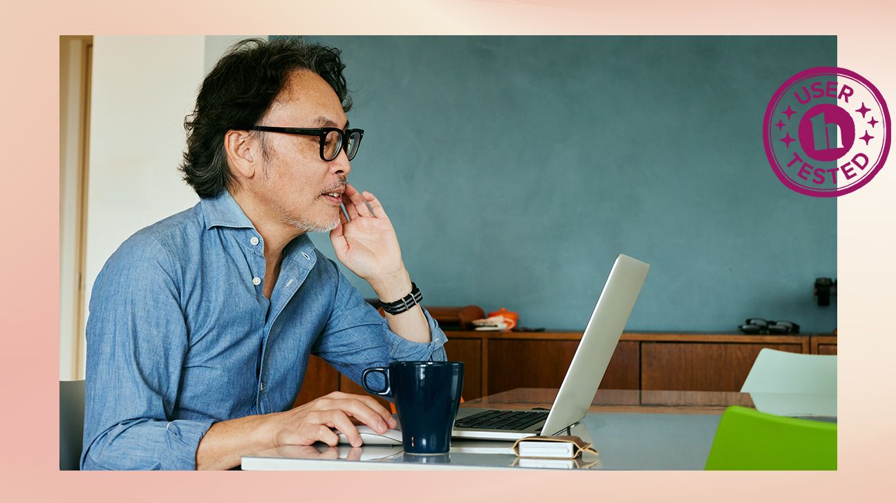 person is sitting at a desk with a laptop and a mug of coffee. Behind them is a chalkboard. They are looking at the laptop with a hand under their chin, and they seem to be moving a mouse around the page.