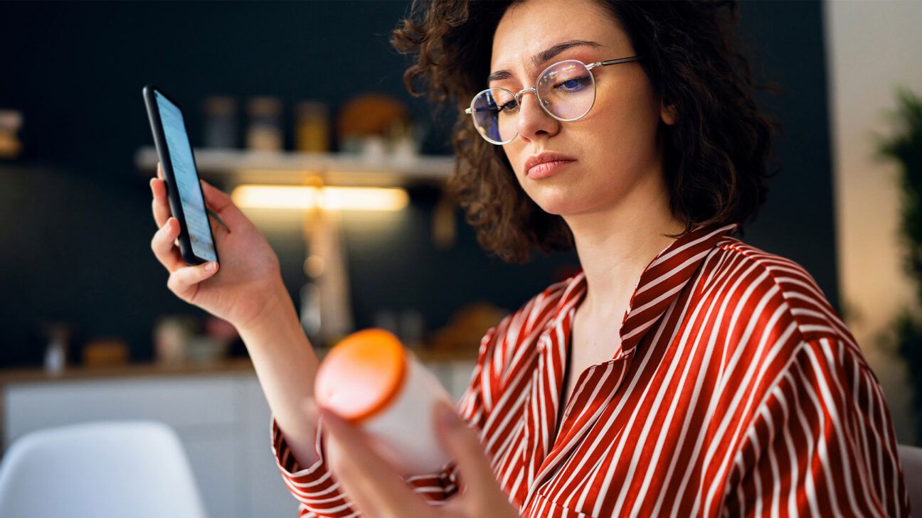 A female holding a bottle of supplements while looking at a smartphone.