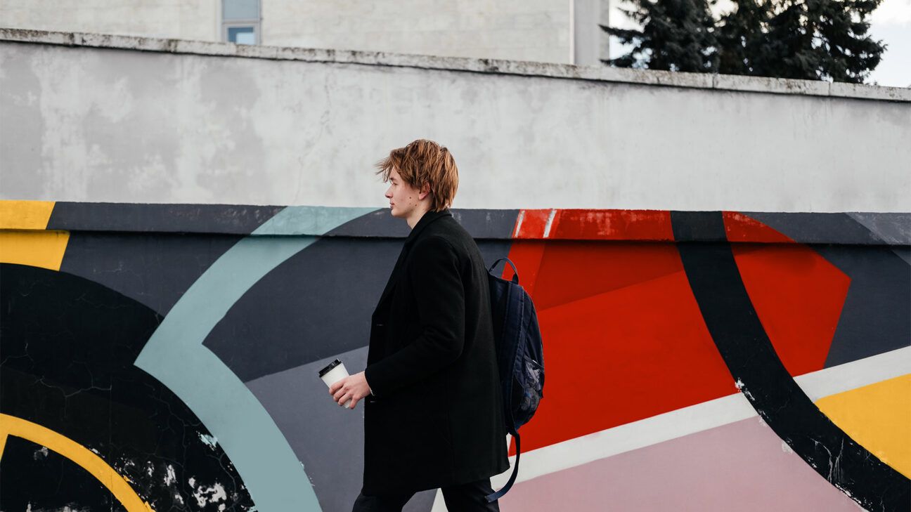 A male wearing all black walking past a painted wall holding a cup of coffee