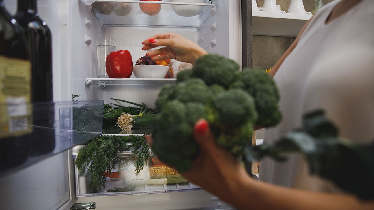 A woman gets broccoli and red pepper out of an open fridge