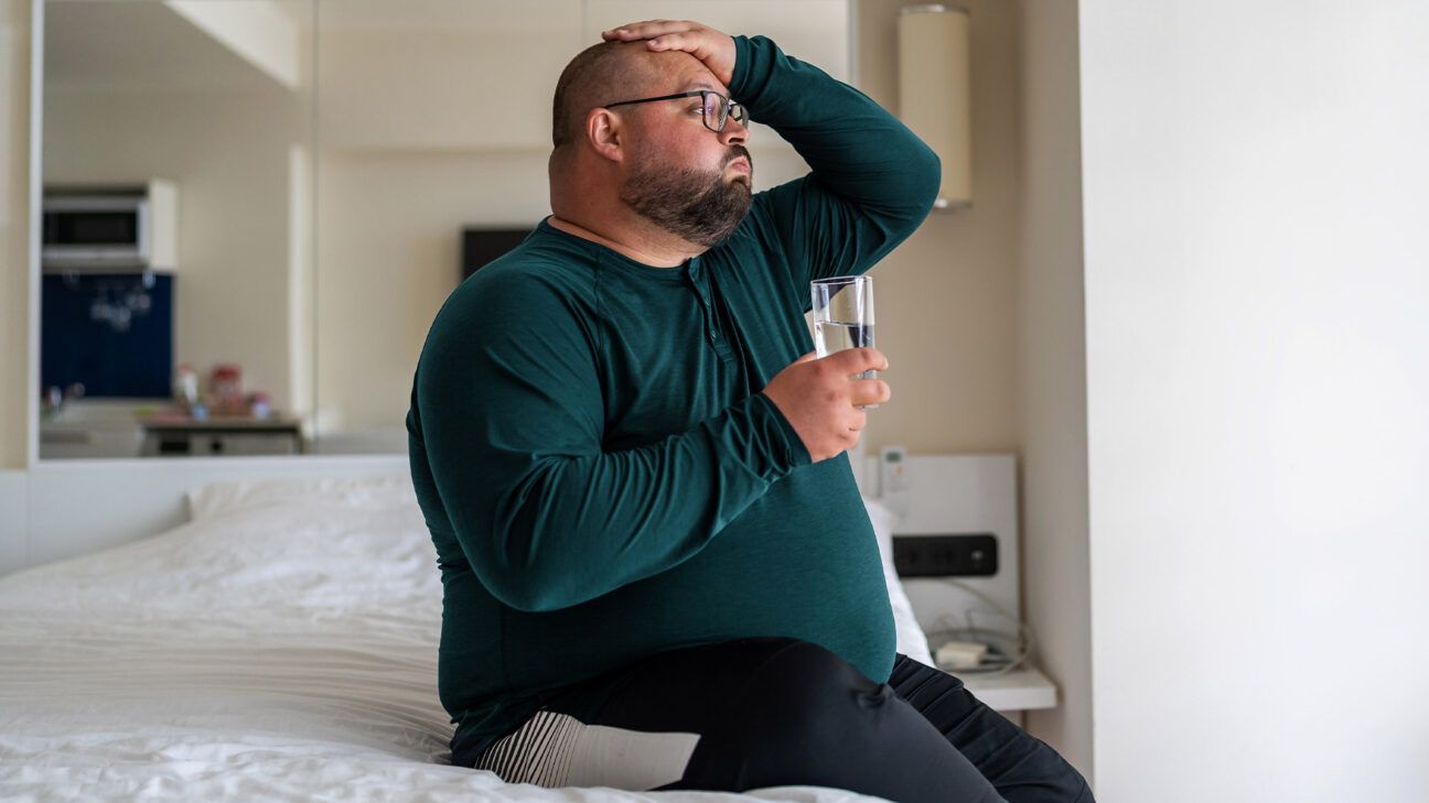 Male sitting on a bed with a glass of water.