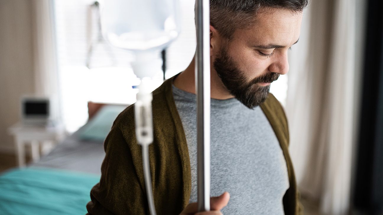 Male in foreground looking slightly downward with eyes closed while standing and holding an infusion pole with an infusion bag attached to it, against a background of a bed