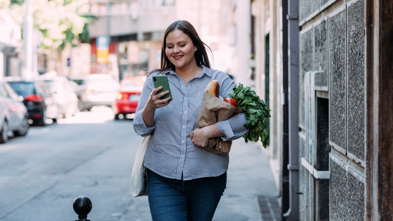 Female holding bag of groceries and cellphone walking down the street