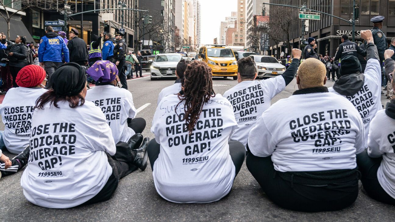 A group of protesters sitting on a street wearing shirts that read, "Close the Medicaid Coverage Gap!"