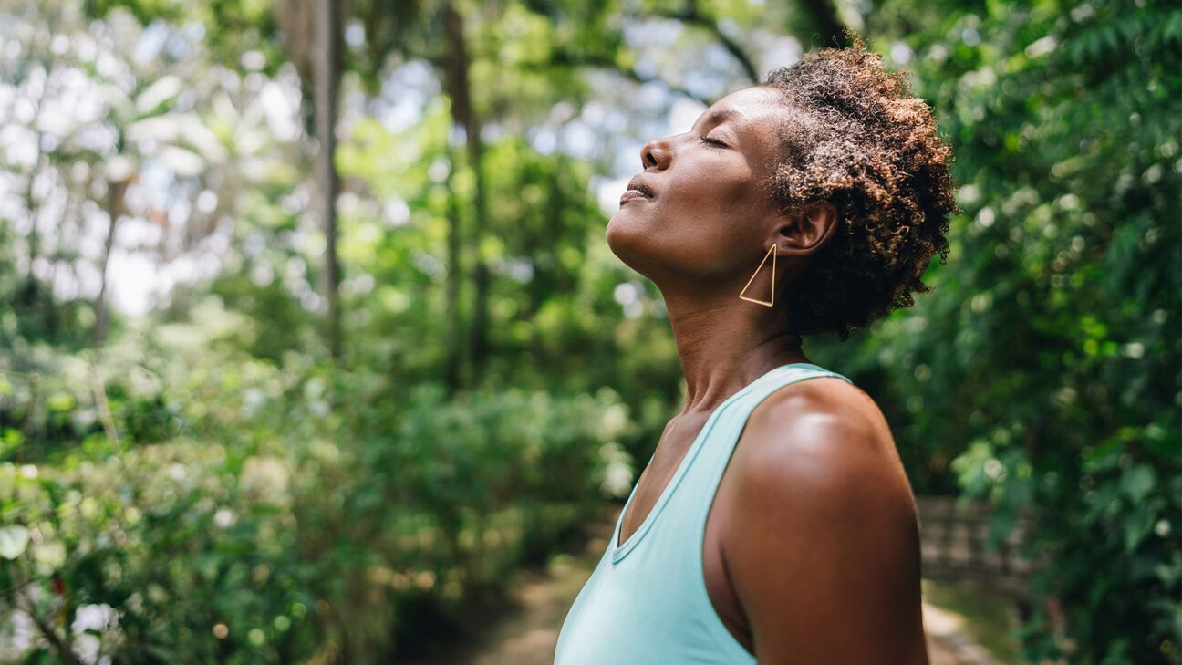 A black woman breathes in fresh air in on a wooded trails with a lot of trees