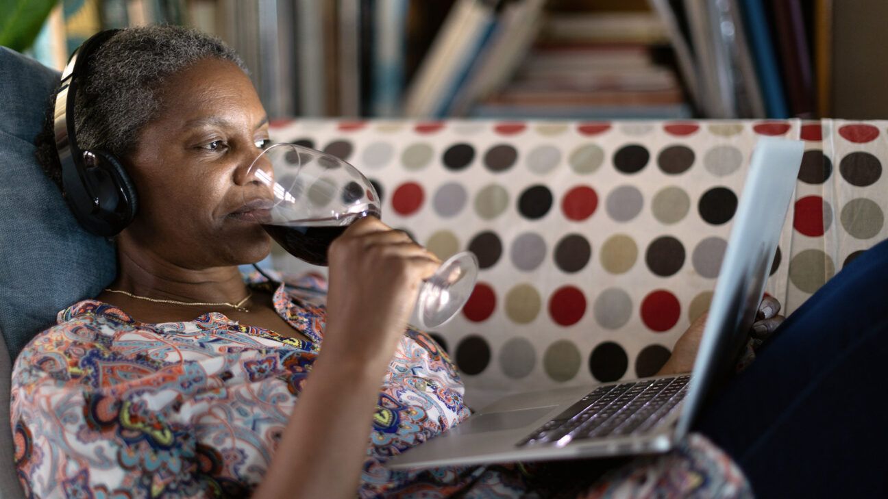 Female drinking wine with headphones and laptop on couch at home