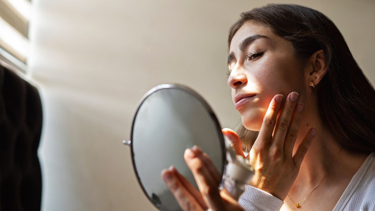 A female looking in a mirror applying lotion to her face.