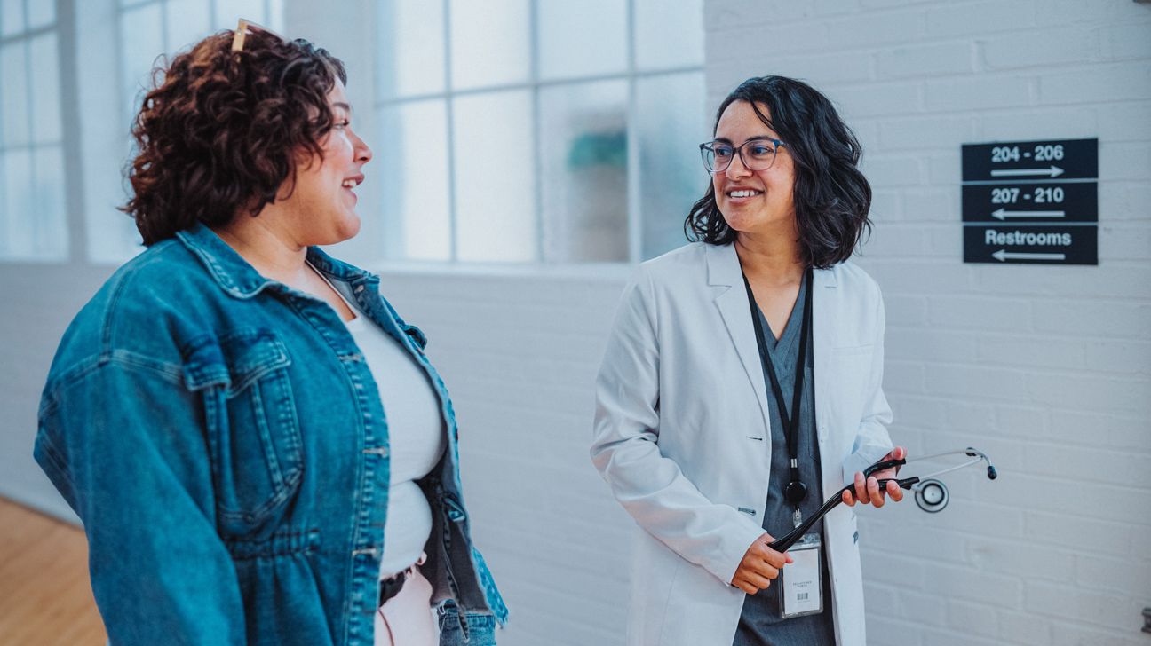 Two women engaged in conversation, one wearing street clothes and the other holding a stethoscope and wearing scrubs and a lab coat