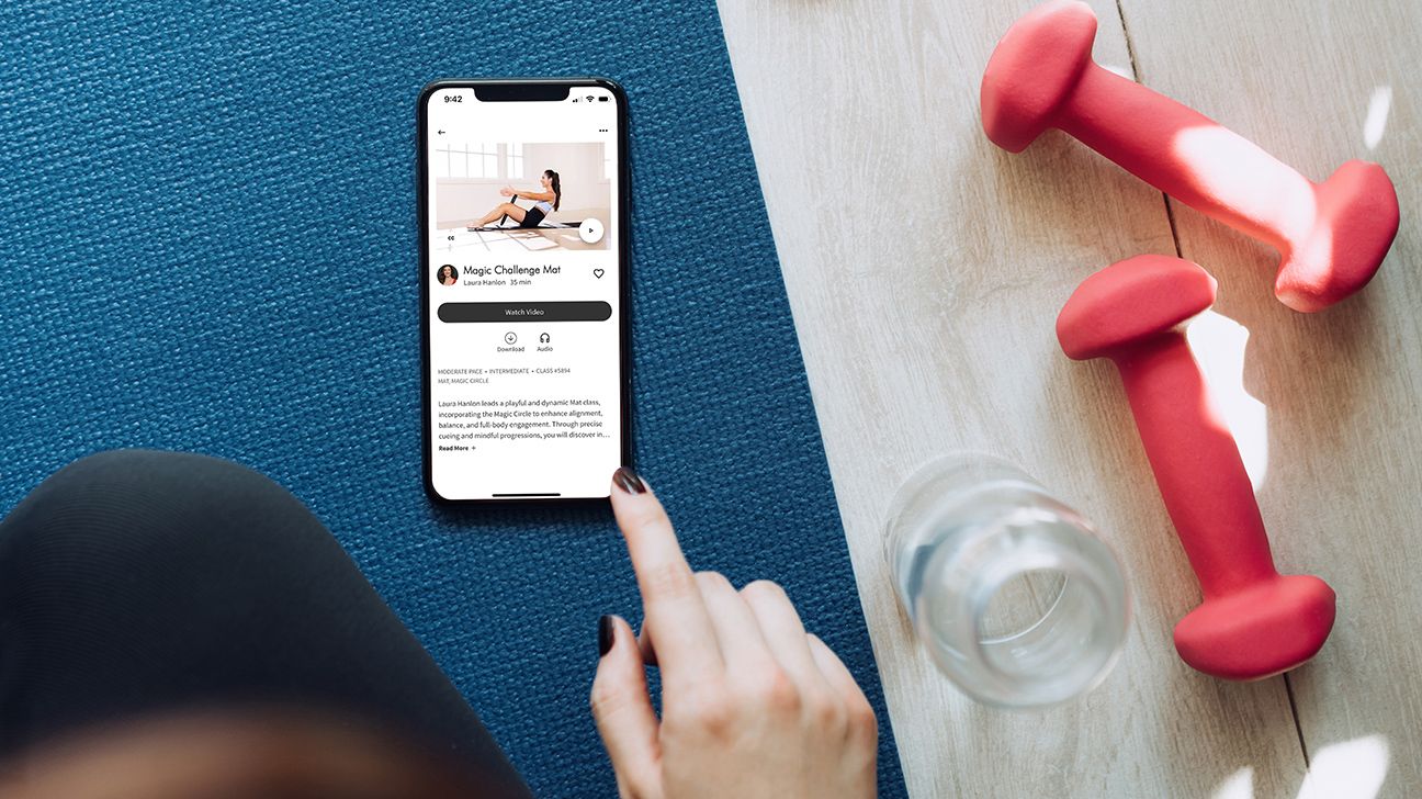 A person scrolling through a Pilates app on their phone, which is resting on a blue mat. There are two red dumbbells and a water bottle positioned to the right. 