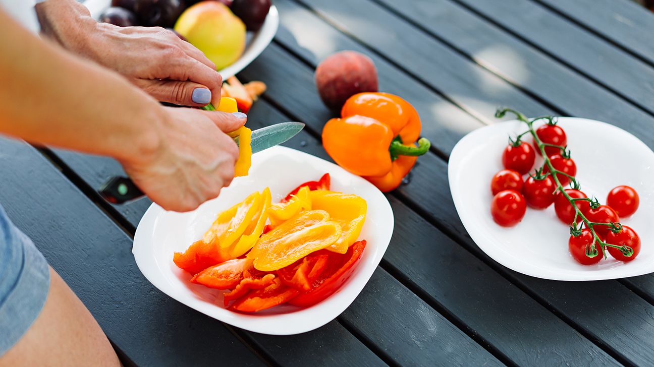 Woman cutting yellow and red peppers in hand onto plate surrounded by more vegetables on a table