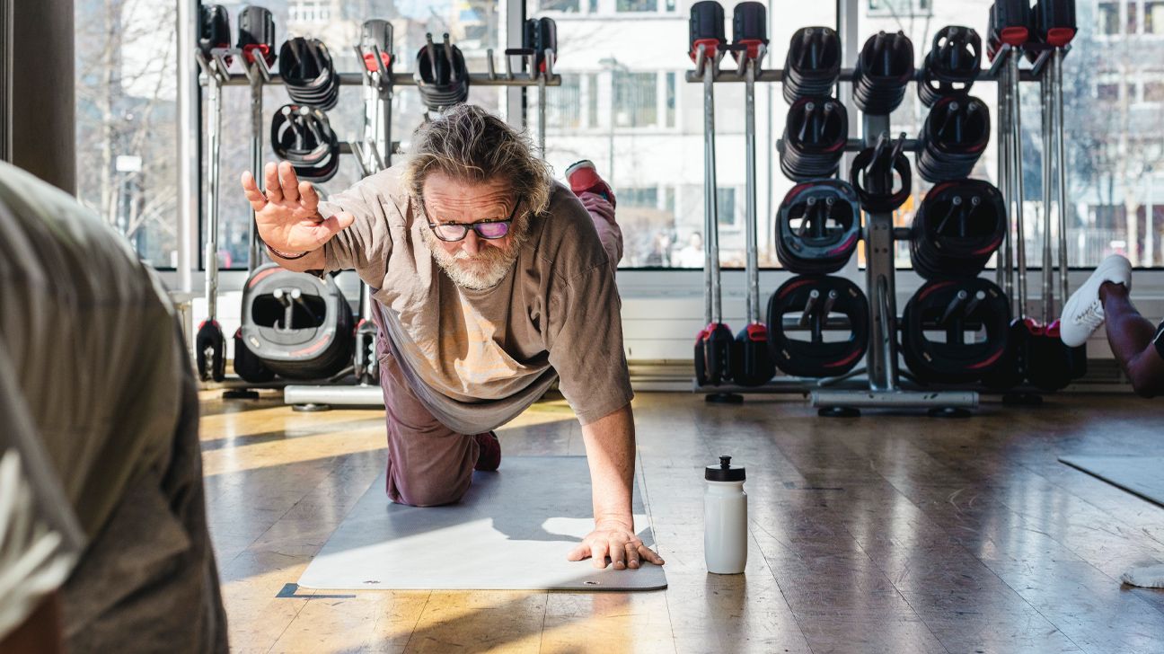 Older adult male stretching on floor exercise mat in a room with weights stacked in the background