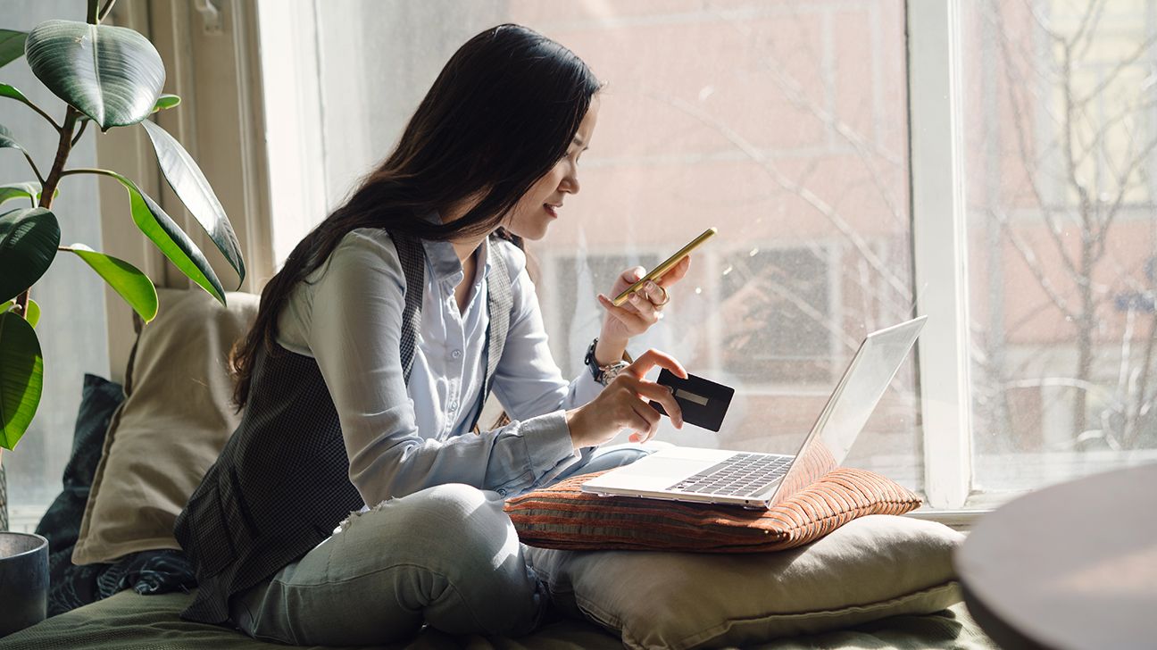 Woman sitting cross-legged, with laptop in front of her, looking at cell phone in one hand, and holding credit card in other hand