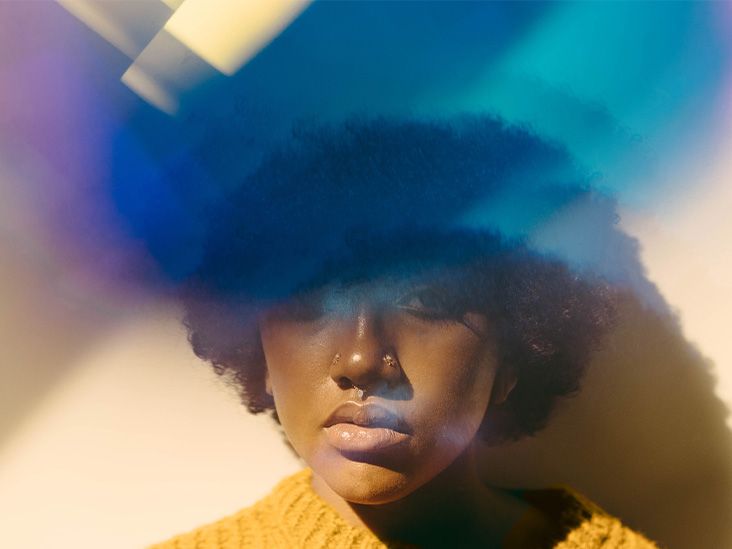 An adult female standing against a wall with purple and blue lighting above her head in a cloud-like manner.