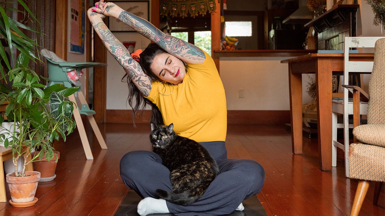 younger white woman with tattoos stretching while in lotus pose on the floor, cat in her lap