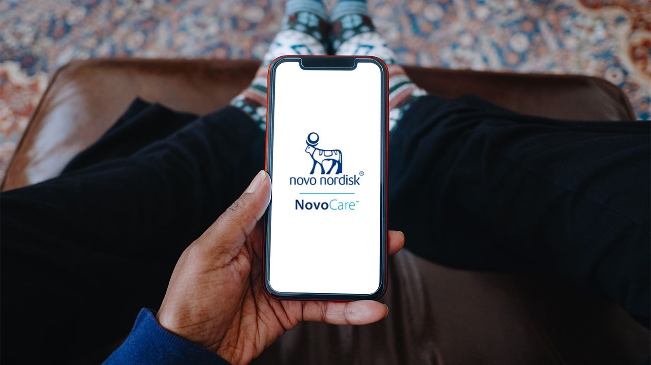 a person is looking down at their phone that is in their left hand. On the phone screen is a white page with the novo nordisk logo in the middle. Underneath that logo is the NovoCare logo