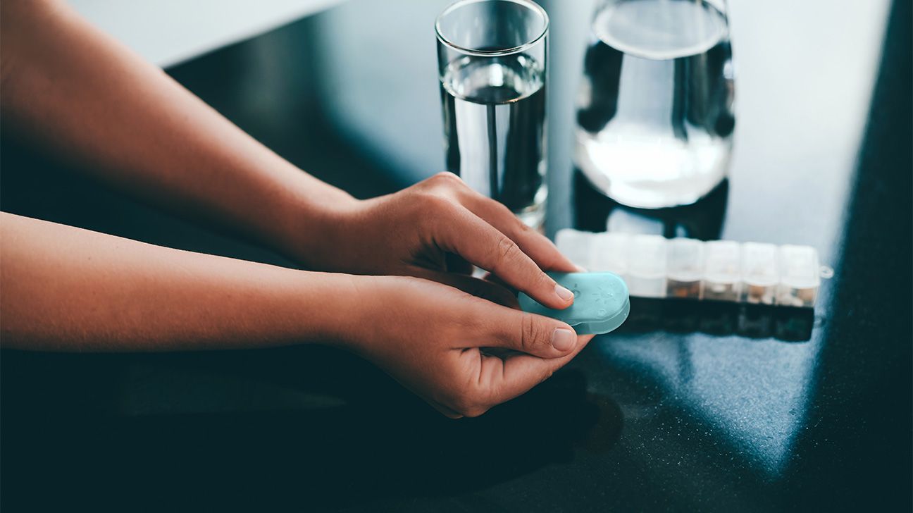 a pair of hands is holding a small, blue pill case. they are about to open it up. To the left of the pair of hands is a full glass of water and a full pitcher of water. Below the glass of water is a larger, week-worth container of medications.