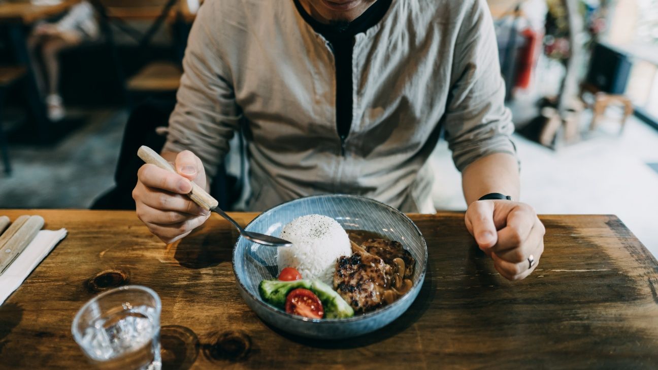 Young Asian man enjoying freshly served beef stew with rice and side salad, sitting at rustic wooden dining table in a restaurant