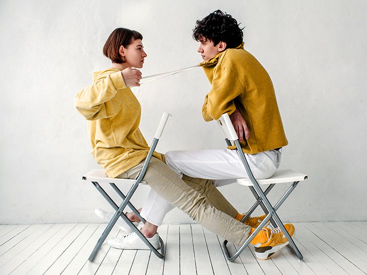 A woman looking exhausted while her partner lounges on the couch, symbolizing the imbalance of emotional labor in relationships.
